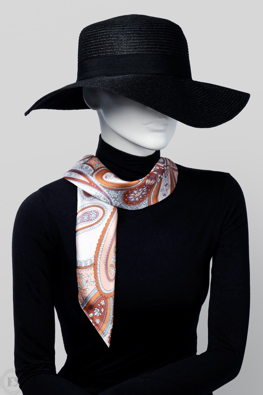 Mannequin i svart hatt och polotröja bär en elegant scarf med paisleymönster i varma nyanser av orange, rosa och vitt, knuten löst runt halsen.