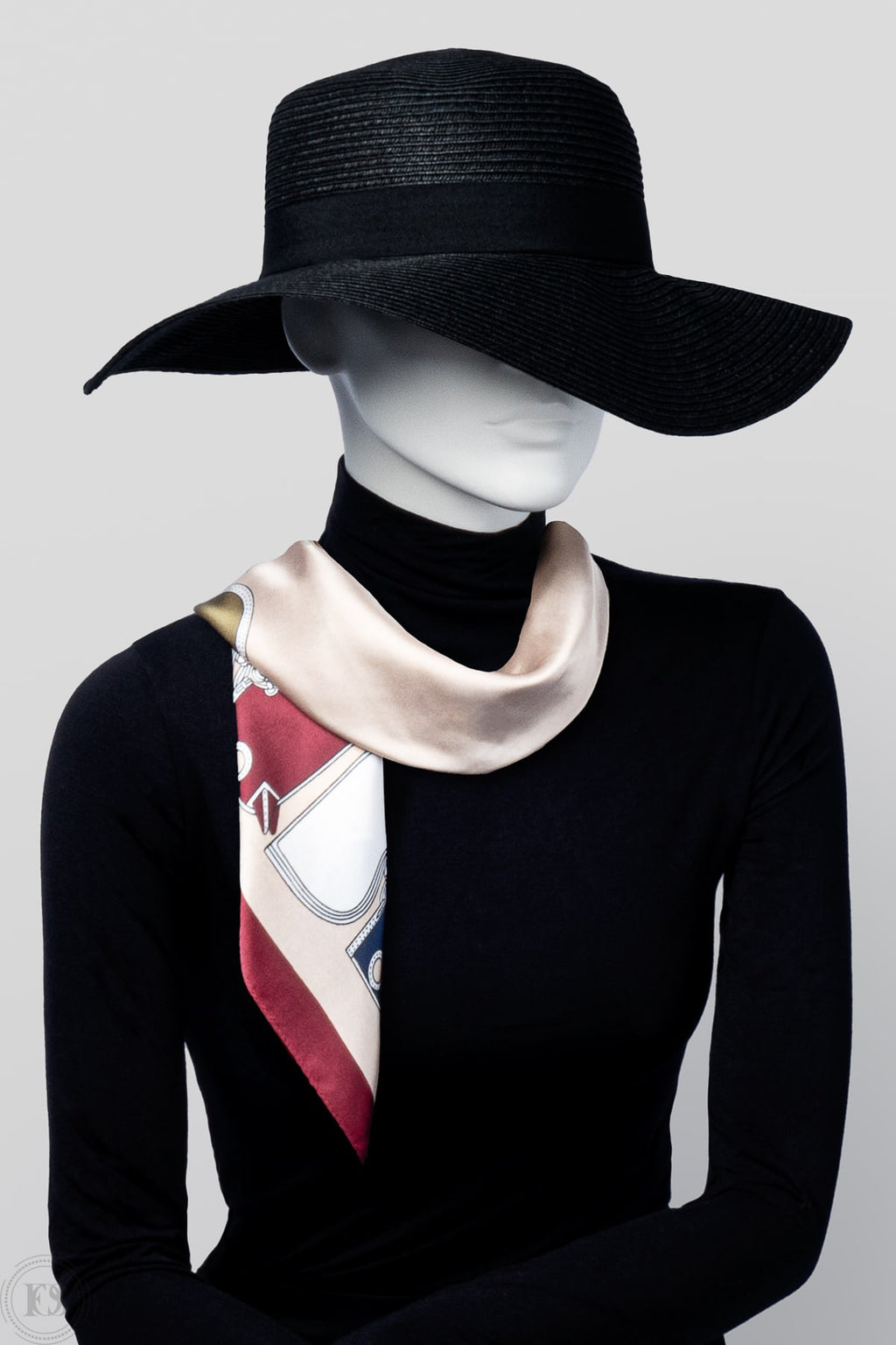 Mannequin i svart hatt och polotröja bär en elegant scarf i beige, rött och vitt med grafiskt mönster. Scarfen är knuten löst runt halsen och har en lyxig sidenlik finish.