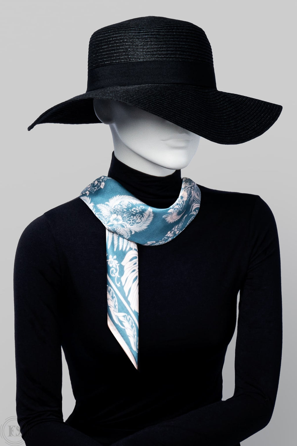 Mannequin i svart hatt och polotröja bär en elegant ljusblå scarf med vitt, romantiskt mönster, knuten löst runt halsen för en sofistikerad look.